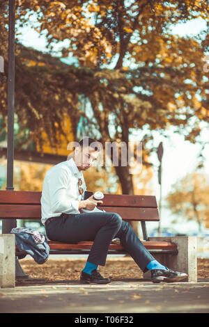 Junge Unternehmer sitzt auf einer Bank im Park, er ist müde und hält er einen Kaffee in Händen zu gehen Stockfoto