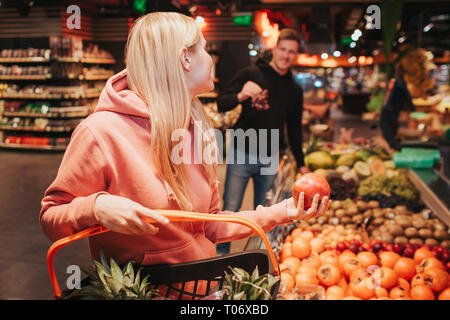 Junges Paar in Lebensmittelgeschäft. Frau pomegranat Halten in der Hand und Blick auf Kerl. Er hat Trauben in die Hände. Lächelnd zu einander. Stockfoto
