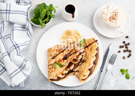 Pfannkuchen mit Banane, Schokolade und Tasse Cappuccino auf konkreten Hintergrund. Ansicht von oben süßes Frühstück essen Stockfoto