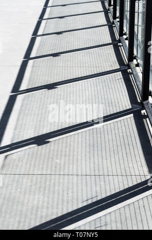 Diagonale Schatten und Drahtgeflecht Muster auf Zement Gehweg aus Metall und Glas panel Zaun Barriere. Stockfoto