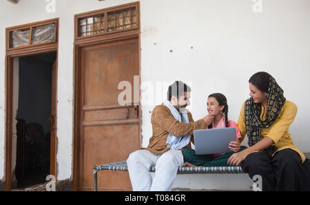 Happy Village Mädchen sitzen mit ihren Eltern auf einem Feldbett mit einem Laptop. Stockfoto
