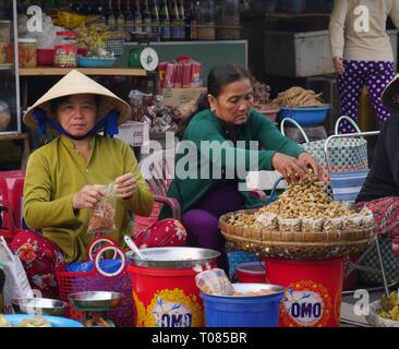 CAN THO, VIETNAM – 2018. MÄRZ: Eine Verkäuferin auf der Straße arrangiert gekochte Erdnüsse, während ein anderer Anbieter Delikatessen für den Verkauf an der Cho an Binh-Marke zubereitet Stockfoto