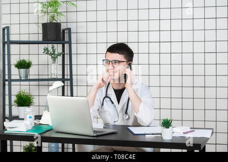Männlicher Arzt über Telefon während der Arbeit am Computer bei Tisch, in der Klinik Stockfoto