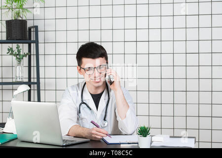 Männlicher Arzt über Telefon während der Arbeit am Computer bei Tisch, in der Klinik Stockfoto