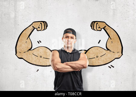 Nahaufnahme Vorderansicht des jungen athletischen Mann mit verschränkten Armen, gegen die Wand, die mit der Ausarbeitung der grossen muskulösen Arme rechts hinter seinem Rücken. Stockfoto