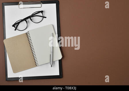 Flach, Ansicht von oben Bürotisch Schreibtisch. Mit leeren Clip Board, Notebook, Brillen, Kugelschreiber auf braunem Hintergrund Arbeitsbereich. Stockfoto