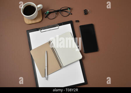 Mockup Arbeitsbereich mit leeren Clip Board, Bürobedarf, Kugelschreiber, Notizblock, Smartphone, Kaffee Tasse und Brillen auf braunem Hintergrund. Flach, Ansicht von oben, Stockfoto