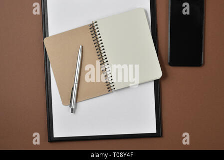Mockup Arbeitsbereich mit leeren Clip Board, Bürobedarf, Kugelschreiber, Notizblock und Smartphone auf braunem Hintergrund. Flach, Ansicht von oben, Kopieren Raum Stockfoto