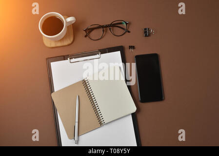 Mockup Arbeitsbereich mit leeren Clip Board, Bürobedarf, Kugelschreiber, Notizblock, Smartphone, Kaffee Tasse und Brillen auf braunem Hintergrund. Flach, Ansicht von oben, Stockfoto