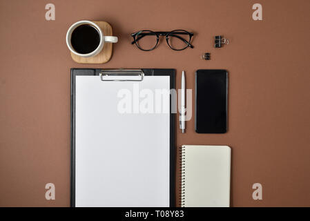 Mockup Arbeitsbereich mit leeren Clip Board, Bürobedarf, Kugelschreiber, Notizblock, Smartphone, Kaffee Tasse und Brillen auf braunem Hintergrund. Flach, Ansicht von oben, Stockfoto