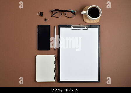 Mockup Arbeitsbereich mit leeren Clip Board, Bürobedarf, Kugelschreiber, Notizblock, Smartphone, Kaffee Tasse und Brillen auf braunem Hintergrund. Flach, Ansicht von oben, Stockfoto