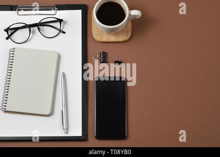 Mockup Arbeitsbereich mit leeren Clip Board, Bürobedarf, pen, Smartphone, Notepad, Kaffee Tasse und Brillen auf braunem Hintergrund. Flach, Ansicht von oben, Stockfoto