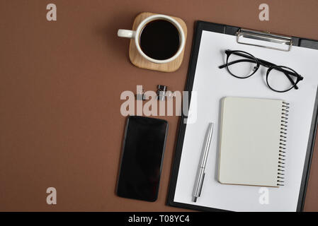 Mockup Arbeitsbereich mit leeren Clip Board, Bürobedarf, pen, Smartphone, Notepad, Kaffee Tasse und Brillen auf braunem Hintergrund. Flach, Ansicht von oben, Stockfoto
