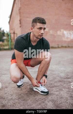 Athlet Mann hockte, Schnürsenkel binden auf Sneakers. Im Sommer auf dem Sportplatz in der Stadt. Trainer Kerl in der Watch tracker Armband nach Stockfoto