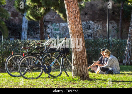 Ein Paar, Leute, die sich unter Bäumen entspannen, Fahrräder parken an der Kiefer, Valencia Turia Gärten, Spanien Fahrrad Stadt Freunde im Park mit Fahrrad, Rasen Stockfoto