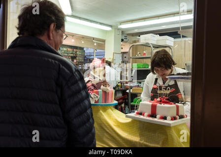 Ein Besucher schaut durch ein Schaufenster, während ein Konditor einen Kuchen auf dem überdachten Golden Cross Market in Oxford, Oxfordshire, Großbritannien, schmückt Stockfoto