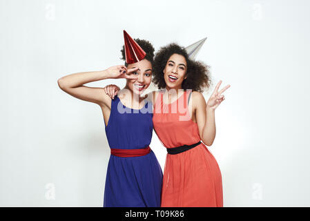 Party Feier. Porträt von zwei positive und junge afro-amerikanische Frauen in schönen Abend Kleider und Hüte zeigen Friedenszeichen und Lachen Stockfoto