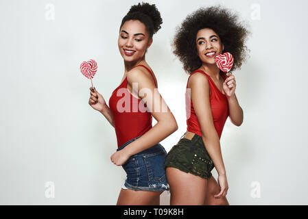 Genießen Sie köstliche Süßigkeit. Zwei attraktive und junge afro-amerikanische Frauen im Sommer Kleidung holding Lutscher geformte Herzen und lächelnd beim Stehen Aga Stockfoto