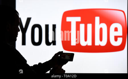 15. März 2019, Schleswig-Holstein, Aukrug-Homfeld: Illustration - ein Mann mit Smartphone steht vor dem Logo der Internetplattform YouTube. Foto: Carsten Rehder/dpa Stockfoto