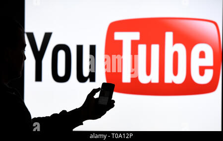 15. März 2019, Schleswig-Holstein, Aukrug-Homfeld: Illustration - ein Mann mit Smartphone steht vor dem Logo der Internetplattform YouTube. Foto: Carsten Rehder/dpa Stockfoto