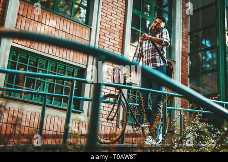 Ein hübscher junger Mann geht in die Stadt mit dem Fahrrad, zu Fuß daneben und sprechen auf dem Smartphone. Stockfoto