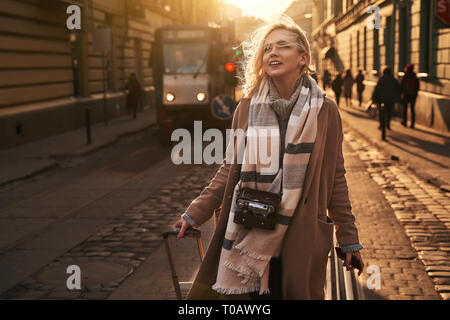 Junge hübsche blonde Frau Tourist mit einer fahrbaren Reisetasche und vintage Film Kamera kommt in eine neue Stadt auf einem sonnigen windigen Abend Stockfoto