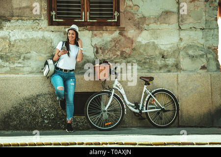 Happy nachdenkliche junge Frau genießt in einem Sommer sonnigen Tag, stützte sich auf ein altes Haus Wand, neben dem Fahrrad mit Blumenkorb. Stockfoto