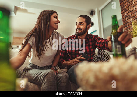 Aufgeregt Paar ist auf der Suche Film auf dem Fernseher zu Hause auf dem Sofa sitzen mit Popcorn und Bier. Sie haben ein tolles Wochenende in Nizza Unternehmen im Innenbereich. Stockfoto