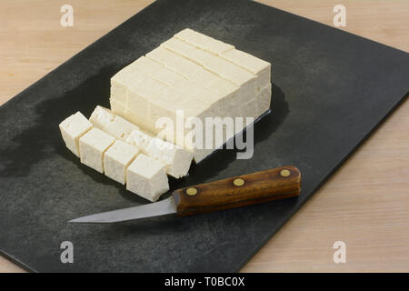 Block von extra festen Tofu in Würfel schneiden auf Schneidebrett mit Messer für das Hinzufügen als Zutat zu Speisen in der Küche vorbereiten Stockfoto