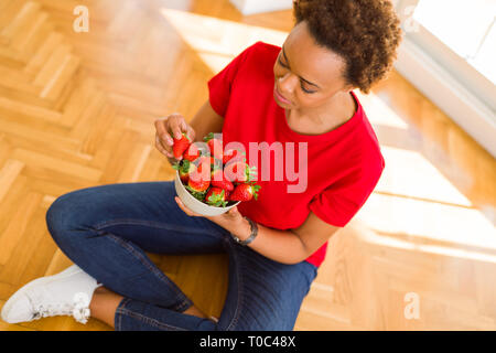 Schöne junge afrikanische Frau mit afro Haar essen frische Erdbeeren sitzen auf dem Boden Stockfoto