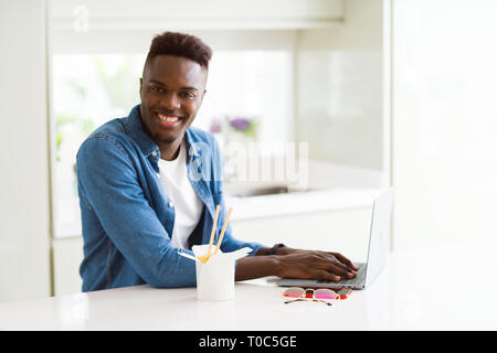 Hübscher junger African Business Mann essen Lieferung asiatische Lebensmittel und Arbeiten mit Computer, genießen Nudeln lächelnd Stockfoto