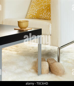 Modernes Wohnzimmer eingerichtet mit komfortablen Sessel mit Kissen, Tisch mit teetasse und Hausschuhe auf weißen Schaffell Teppich. Entspannt zu Hause. Stockfoto
