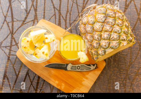 Blick von oben auf die in Scheiben geschnittene Ananas gehalten auf einer hölzernen Tisch neben einem Glas mit Ananas Saft gefüllt und mit einem Glasgefäß mit ananasscheiben und ein Messer Kep Stockfoto