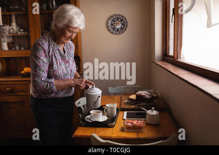 Active Senior Frau Kaffee in einer Schale am Esstisch in der Küche zu Hause gießen Stockfoto