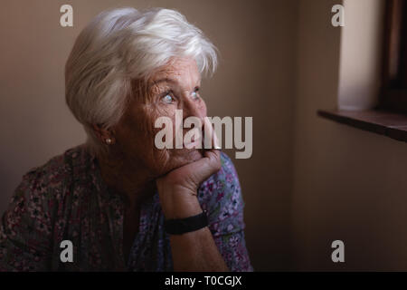 Nachdenklich Active Senior Frau mit der Hand am Kinn durch das Fenster in der Küche zu Hause suchen Stockfoto