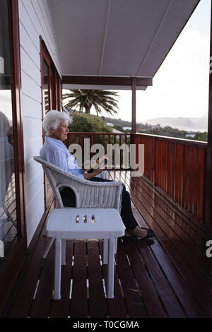 Active Senior Frau liest ein Buch, während auf Stuhl auf dem Balkon zu Hause sitzen Stockfoto