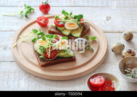 Gesunde Sandwiches mit Avocado, Tomaten, Wachteleier und Sonnenblumen micro Grünen (Sprossen) auf weißem Holz- Hintergrund Stockfoto