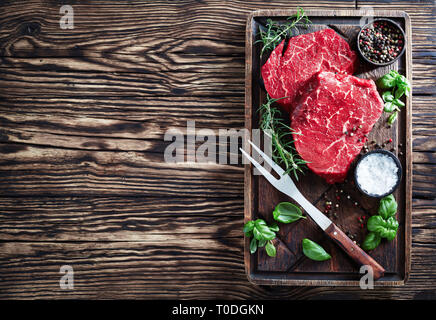 Ansicht von oben von rohem Rindfleisch Steaks mit Pfefferkörnern, frischem Basilikum, Rosmarin, Salz und Gabel auf eine unhöfliche Schneidbrett auf einem alten rustikalen Holztisch, Stockfoto