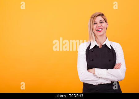 Schöne junge blonde Frau in den Barkeeper gleichmäßig über der Hintergrund gelb gekleidet. Gekreuzte Arme. Weißes T-Shirt. Stockfoto