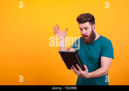 Gut aussehender bärtiger Mann im grünen T-Shirt auf der Suche nach Tablet auf gelben Hintergrund schockiert. Coole hipster Mann. Stockfoto