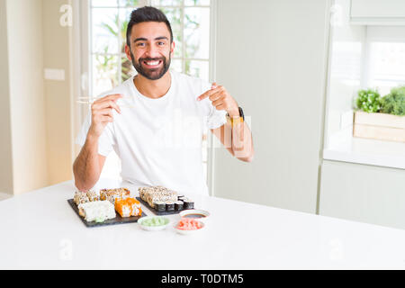 Stattliche Hispanic Mann essen asiatische Sushi mit Stäbchen mit Überraschung Gesicht zeigenden Finger zu sich selbst Stockfoto