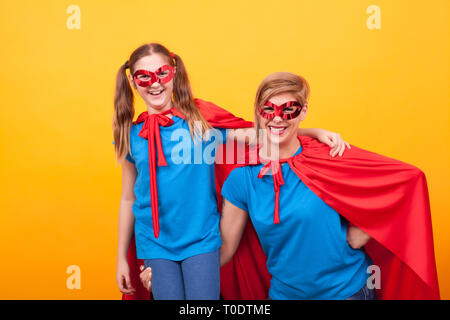 Superhelden Mutter ihr kleines Mädchen Halten auf ihrem Arm auf gelben Hintergrund. Mutter und Tochter lächelnd. Parenting. Rote Maske. Roten Cape. Stockfoto