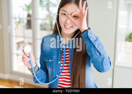 Schöne asiatische Frau prüfen health care mit Stethoskop mit glücklichen Gesicht lächelnd tun ok Zeichen mit der Hand auf Auge schauen durch die Finger Stockfoto