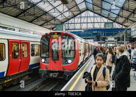 Menschen, die auf dem Bahnsteig warten, während ein Zug der District Line in die U-Bahnstation Earls Court, London, Großbritannien, fährt Stockfoto