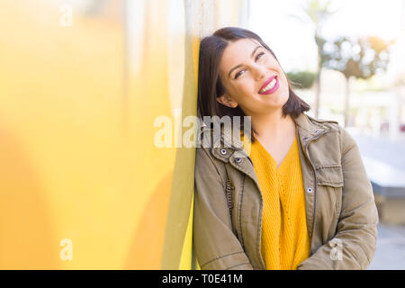 Schöne junge Frau lächelte zuversichtlich und fröhlich lehnte sich auf gelbe Wand, gehen auf die Straße der Stadt an einem sonnigen Tag Stockfoto