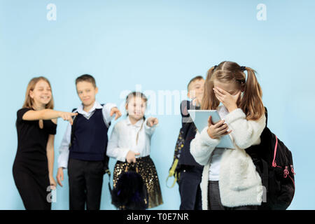 Kleine Mädchen, die alleine stehen und Leiden ein Akt von Mobbing, während Kinder verspotten. Traurige junge Schülerin sitzen auf Studio vor blauem Hintergrund. Stockfoto