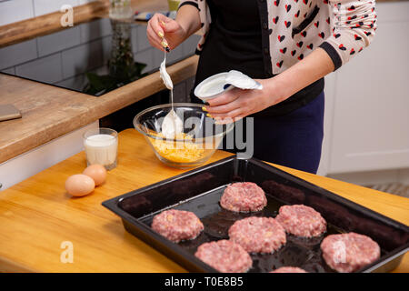 Frau giesst saure Sahne, geriebenen Käse auf moderne Küche Hintergrund. Frauen Kochen das Fleisch zum Braten im Ofen Stockfoto