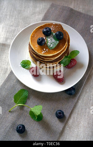 Hausgemachte Pfannkuchen mit Waldfrüchten Beeren auf weißer Tisch. Frische Pfannkuchen auf Platte Tischdecke. Blaubeeren, Himbeeren, Minze auf Pfannkuchen. Stockfoto