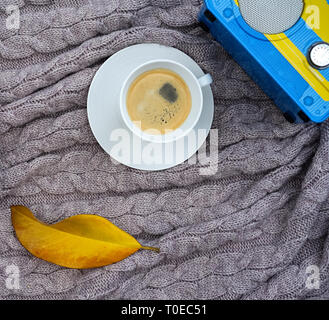 Weiße Tasse Kaffee und gelb blau Funkempfänger und gefallenen gelben Blatt auf warme Wolldecke Grau Pink Plaid. Gestrickte textile mit einem Geflecht Muster. Herbst Winter Konzept von Komfort und Entspannung. Stockfoto