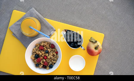 Gesundes Frühstück mit Müsli, griechischer Joghurt, Apfel, Beeren und frischem Orangensaft auf gelb tischdecke Schneidebrett auf grauem Hintergrund. Flach, Gesundheitswesen Konzept. Happy morgen Zeit. Stockfoto
